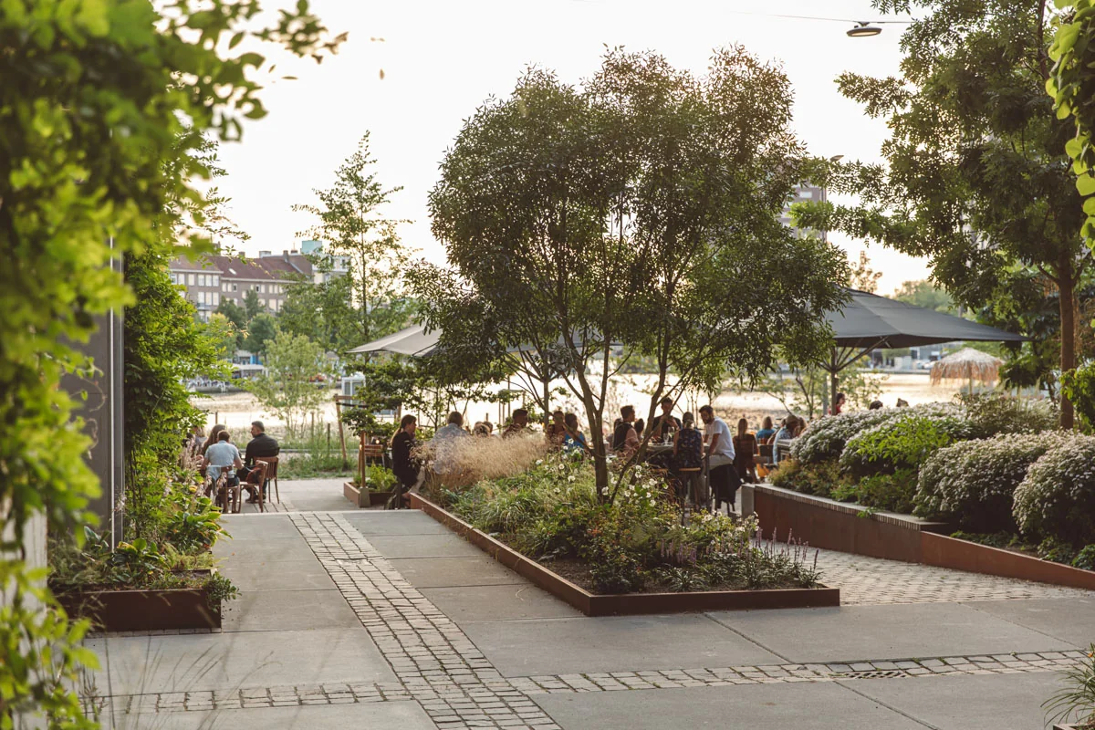 Terras aan de Karl-Weisbardstraat met mensen die gezellig buiten zitten tussen weelderig groen en bloemen.